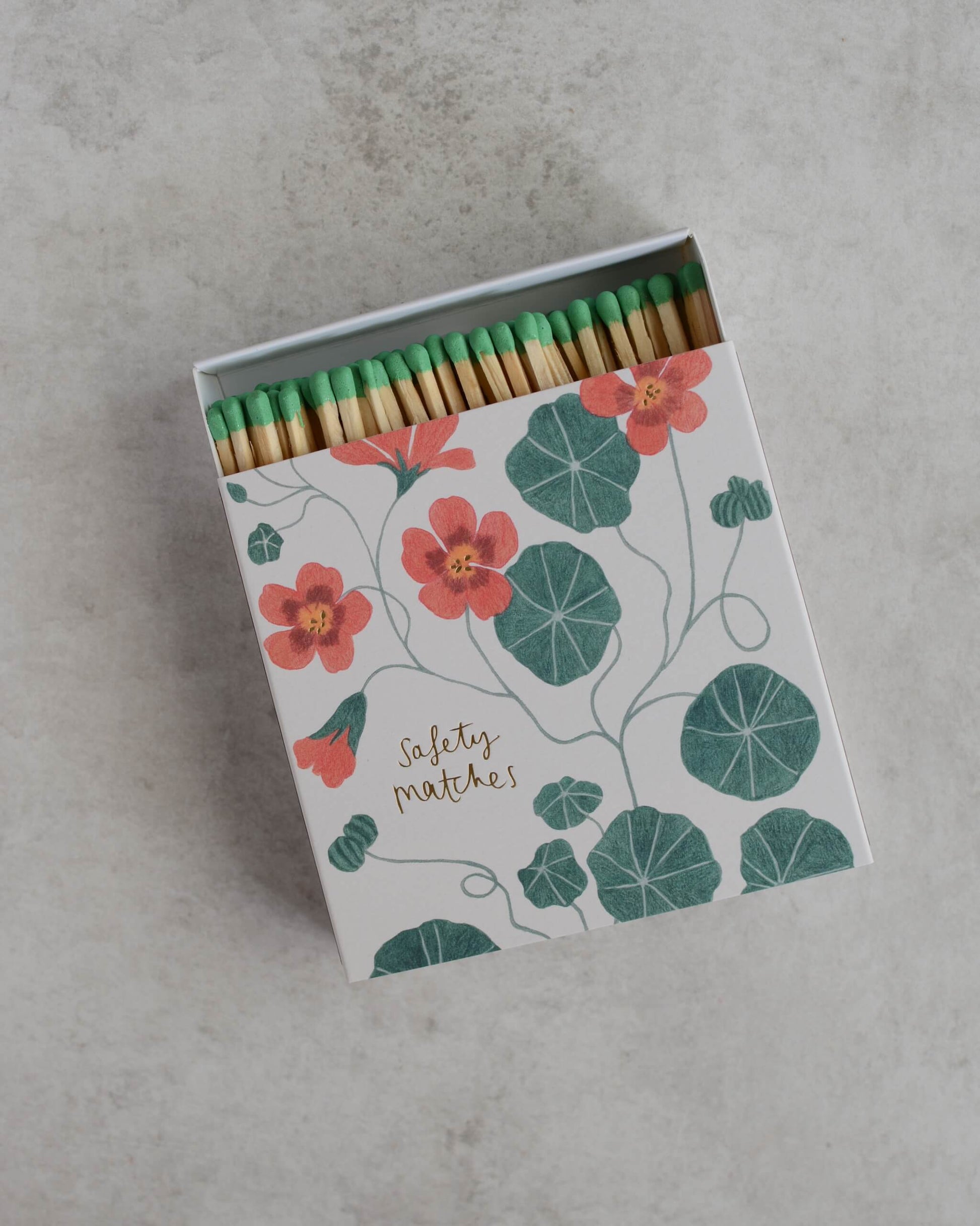 A decorative matchbox filled with safety matches, photographed from above on a light grey surface. The box features a botanical illustration with coral-pink nasturtium flowers and green leaves, with a gold script reading 'safety matches'.