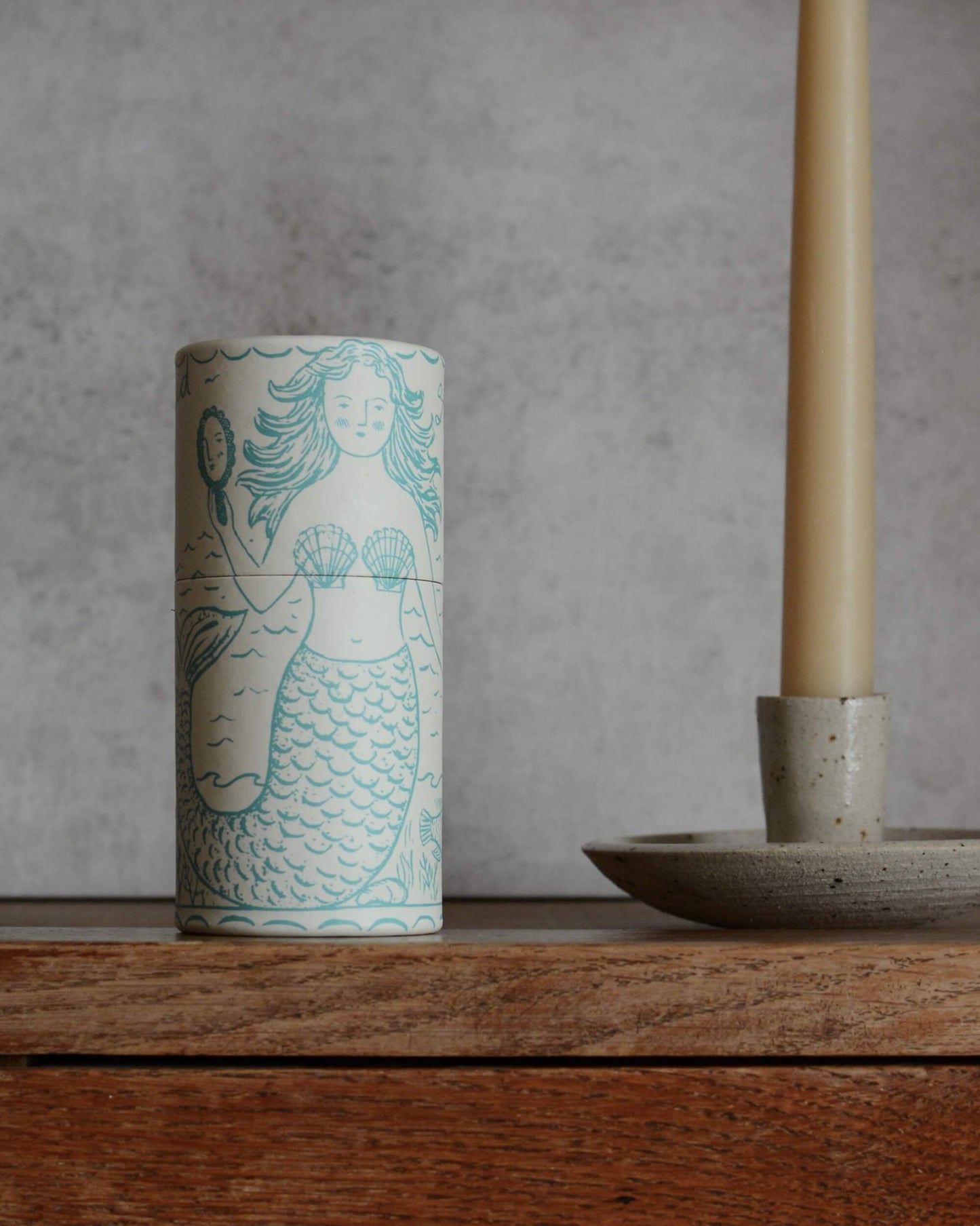 A cylindrical white matchbox with a turquoise mermaid illustration, set on a rustic wooden surface next to a ceramic candlestick holder.