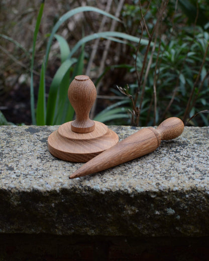 An oak seed dibber and pot tamper, shown on a garden wall with plants behind.