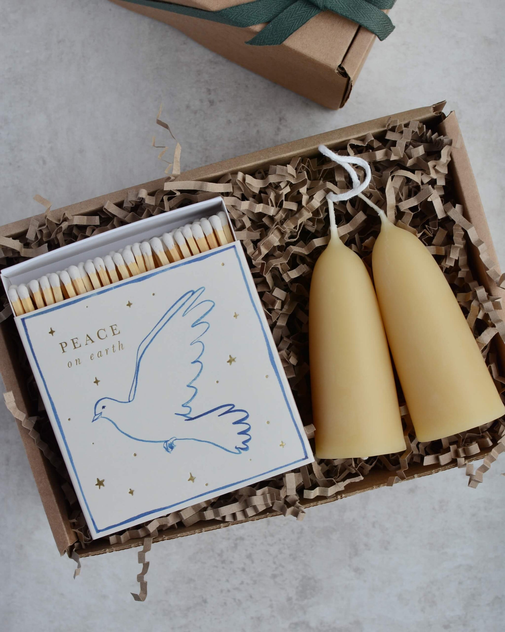 A pair of stubby beeswax candles and a box of matches with an illustration of a peace dove with gold lettering.