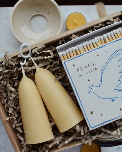 A pair of stubby beeswax candles and a box of matches with an illustration of a peace dove, alongside ceramics and beeswax tealights.