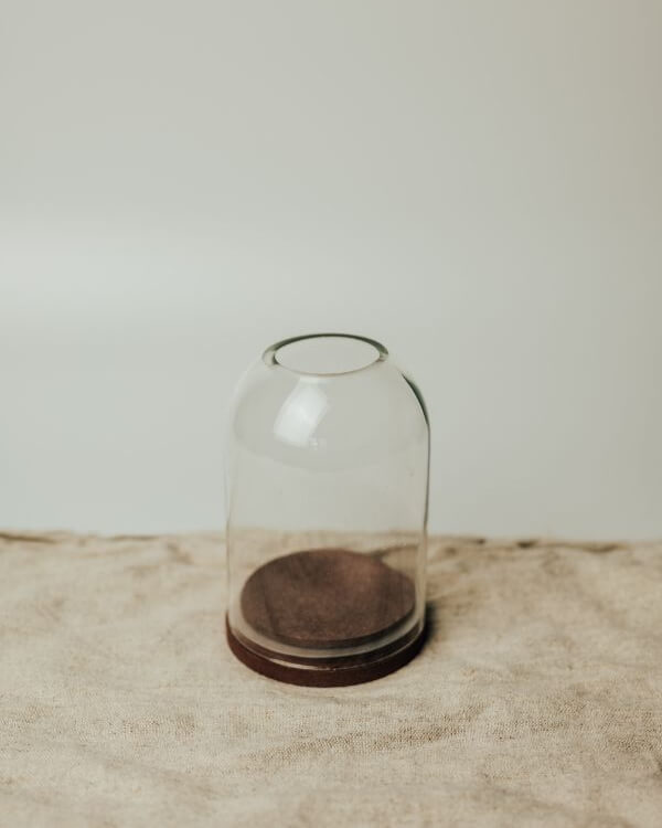 A clear glass cloche candle dome with a dark brown wooden base, set on a textured beige fabric surface.