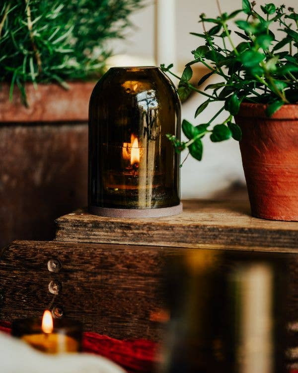 An olive green glass jar candle lantern with a lit candle inside, styled on a rustic wooden surface next to potted green plants.