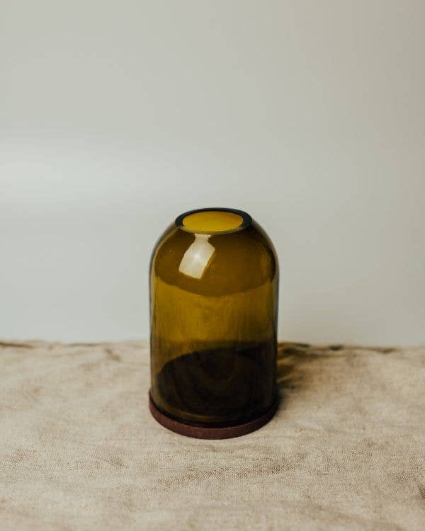 An olive green glass dome candle holder with a dark brown wooden base, set on a textured beige fabric surface.