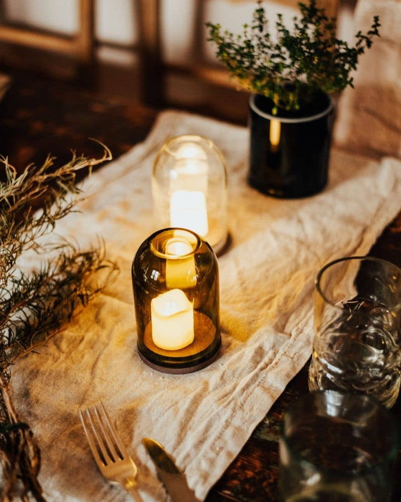 A warm, rustic tabletop setting featuring glass dome candle holders, alongside other decorative on a linen runner.