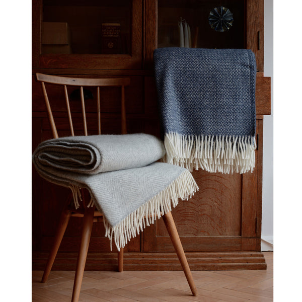 A mid blue Tweedmill windmill wool throw with an ivory fringe, shown infront of a wooden cabinet.