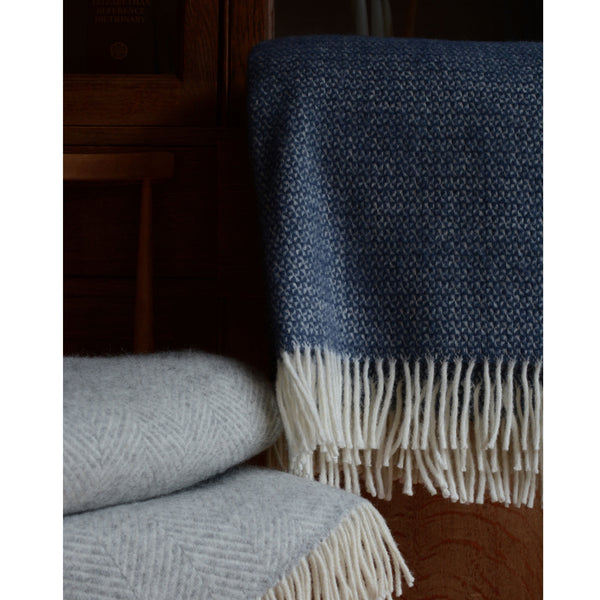 A close up of a mid blue Tweedmill windmill wool throw with an ivory fringe and a folded grey blanket.