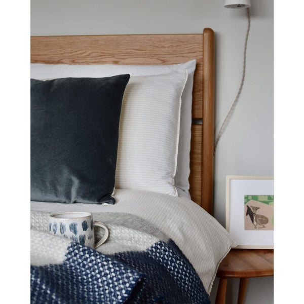 Tweedmill light grey and navy cross weave wool throw, draped on the edge of a bed, with a coffee cup and velvet cushion.
