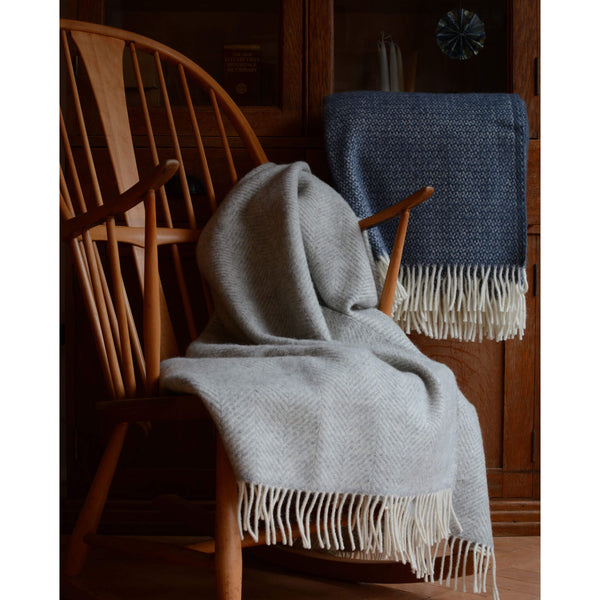 Tweedmill pale grey wool throw with a herringbone pattern and ivory fringe, shown draped over an Ercol rocking chair.