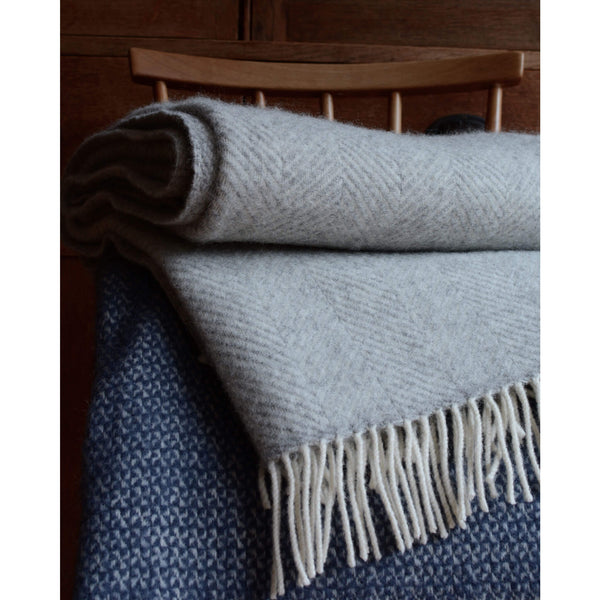 Tweedmill pale grey wool throw with a herringbone pattern and ivory fringe, folded on a chair, on top of a blue windmill pattern blanket.
