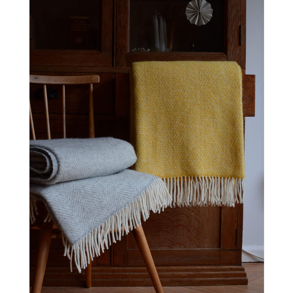 Tweedmill pale grey wool throw with a herringbone pattern and ivory fringe, shown in front of a wooden cabinet and next to a bright yellow blanket.