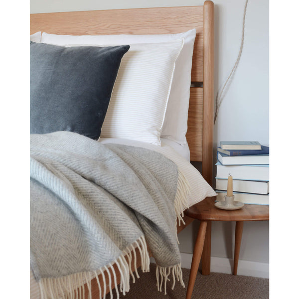 Tweedmill pale grey wool throw with a herringbone pattern and ivory fringe, draped over the edge of a wooden bed.