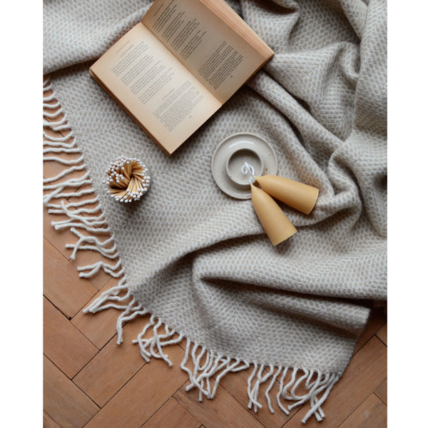 An overhead shot of a Tweedmill oatmeal beehive wool throw laid out on a parquet wooden floor, with candles, candle holder, matches and an open book.