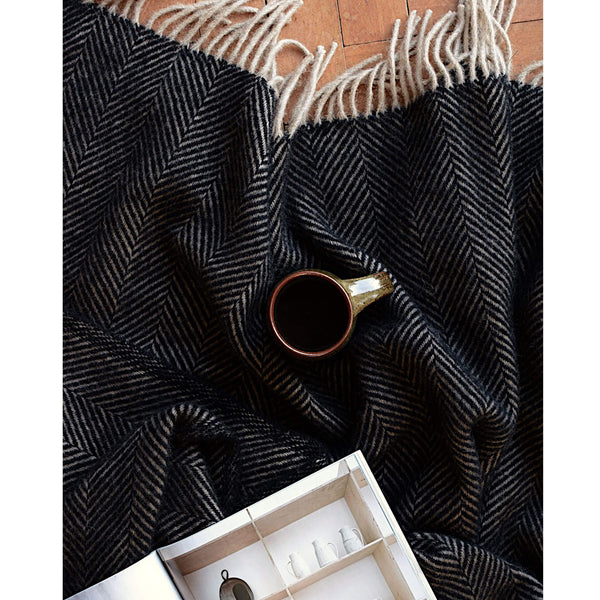 A dark brown wool throw with a herringbone pattern and fringe, shown on a parquet floor with a coffee cup and open magazine.