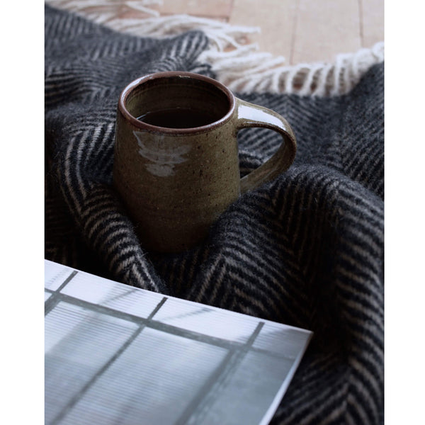 A close up of a dark brown wool throw with a herringbone pattern and fringe, shown with a coffee cup and open magazine.