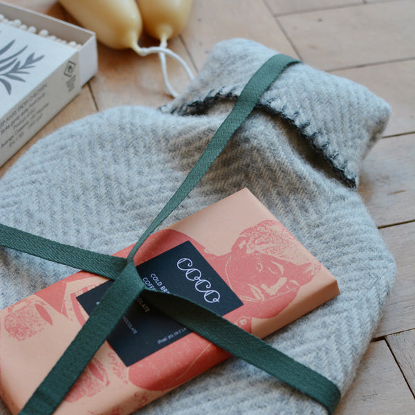 A grey wool hot water bottle with a herringbone pattern, shown with a chocolate bar and wrapped in green ribbon.
