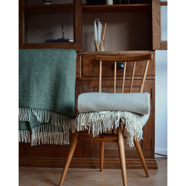 An olive green Tweedmill herringbone wool throw, shown infront of a vintage wooden school cabinet, next to another blanket on a chair.