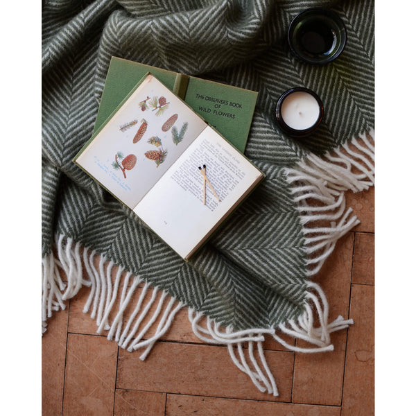 An overhead shot of an olive green Tweedmill herringbone wool blanket with a selection of open books and candle.