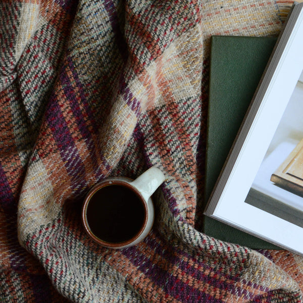 A close up of a recycled wool Tweedmill blanket, with a coffee cup and book on top.