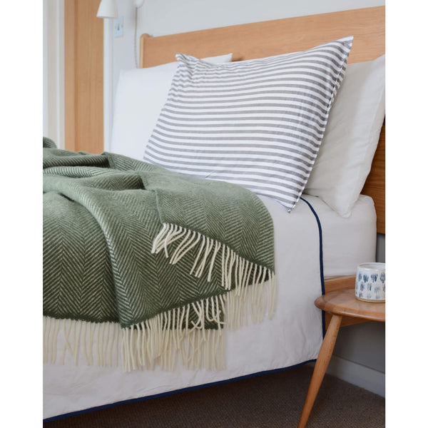 Sage green blanket with an ivory fringe, shown draped across a bed.