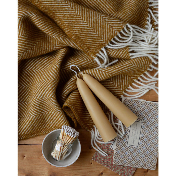 An overhead shot of a mustard yellow herringbone wool throw with an ivory fringe, shown next to a pair of beeswax candles and some music books.
