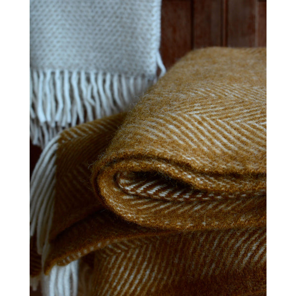 A close up of a folded mustard yellow herringbone wool throw with an ivory fringe.