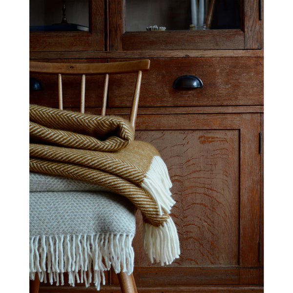 A mustard yellow herringbone wool throw with an ivory fringe, shown folded on top of a wooden chair.
