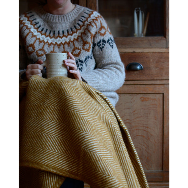 A woman sat on a chair, with a mustard yellow herringbone wool throw across her lap.
