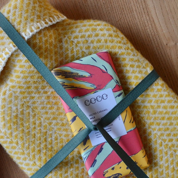 A bright yellow wool hot water bottle with a beehive pattern with a brightly coloured chocolate bar.